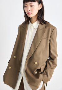 Hnědý dvouřadý blazer s velkými krémovými knoflíky, ostrými klopy a hladkým povrchem, přeložený přes světle krémovou košili.