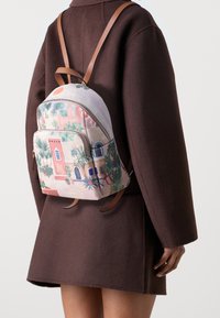 Persona che indossa un cappotto marrone scuro con uno zaino piccolo caratterizzato da illustrazioni colorate di edifici e piante sulla parte posteriore.