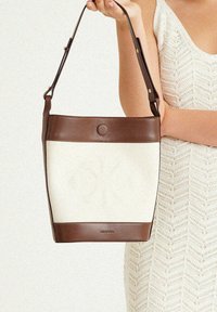 Braune Leder-Eimer-Tasche mit einem cremefarbenen Canvas-Korpus, ausgestattet mit einem runden Druckverschluss und einem verstellbaren Tragegurt. Strukturiert Oberfläche und minimalistisches Design.