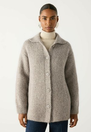 Kvinde iført en beige, blød cardigan med knapper over en cremefarvet rullekravesweater og mørkeblå jeans, stående mod en ensfarvet baggrund.