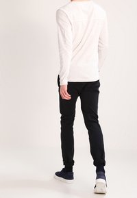 Långärmad, ljusbeige skjorta med texturerad tyg, i kombination med mörka slim-fit-byxor och marinblå sneakers, sedd bakifrån.