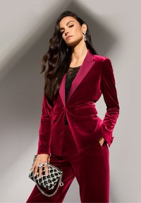Vrouw in een rood fluwelen pak met een zwart kanten top, die een zwarte clutch met zilveren kettingversiering vasthoudt en grote kristallen oorbellen draagt.
