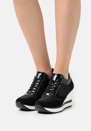 Jambes portant des baskets compensées noires avec des lacets noirs, des accents en cuir verni, et des semelles compensées blanches sur un fond blanc.