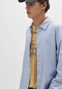 Lichtblauwe gestreepte button-up shirt met witte knopen, gedragen over een beige graphic t-shirt. Inclusief gelaagde ketting; casual ontwerp.