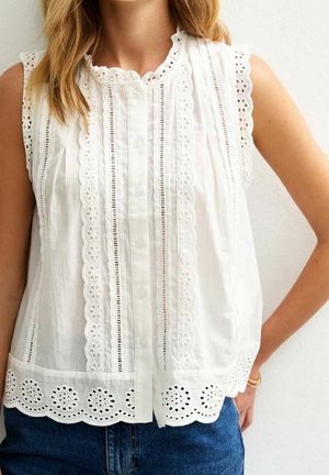 Kvinde iført en ærmeløs hvid bluse med blomstrede øjenhulsblonde-detaljer, bølgede kanter og knapper foran, kombineret med blå jeans.