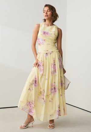 Femme vêtue d'une robe maxi jaune à fleurs sans manches avec un corsage froncé, tenant une pochette beige, et portant des sandales blanches à talons et à brides.