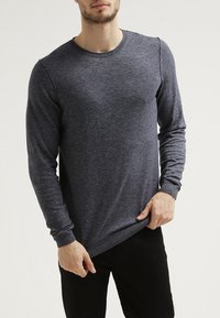 Mann in einem eng anliegenden, dunkelgrauen Langarmpullover und schwarzen Hosen, der mit seiner linken Hand den Saum des Pullovers anpasst.