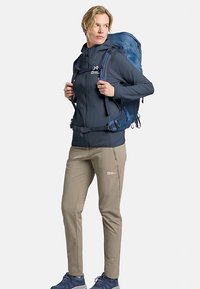 Veste polaire bleu marine, pantalon beige, et sac à dos bleu avec bretelles ajustables, comprenant une poche frontale zippée et un design texturé.