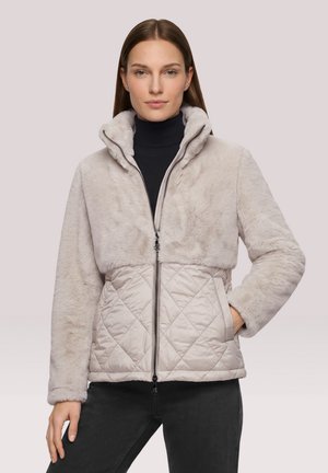 Femme portant une veste beige à fermeture éclair avec une moitié inférieure matelassée et une moitié supérieure douce et duveteuse, debout avec une main dans la poche.