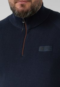 Marineblauwe trui met ribbelstructuur, halve rits en een rubberen logo-label op de borst. Voorzien van een hoge kraag.