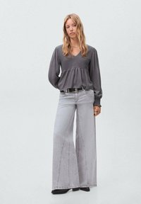 Femme debout portant un chemisier gris à manches longues avec taille smockée et un jean gris clair à jambes larges, avec des bottes noires, sur un fond clair.