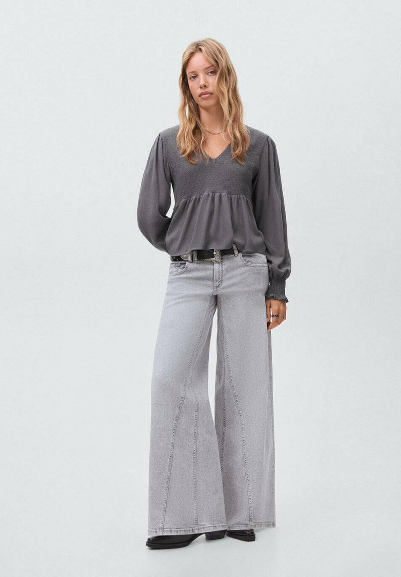 Femme debout portant un chemisier gris à manches longues avec taille smockée et un jean gris clair à jambes larges, avec des bottes noires, sur un fond clair.
