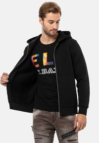 Haut à capuche noir zippé avec une texture douce, doté d'une poche kangourou et de poignets côtelés, porté sur un t-shirt noir avec des lettres colorées.