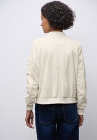 Veste bomber couleur crème avec une texture côtelée, des poignets élastiques et une taille élastique. Portée sur un jean bleu foncé, vue de dos.