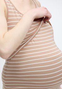 Έγκυος σηκώνει ριγωτό μπεζ τοπ, αποκαλύπτοντας το πάνω μέρος της κοιλιάς και το χέρι σε απλό φωτεινό φόντο.