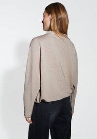 Donna con capelli lisci castano chiaro, che indossa un maglione beige a coste e jeans neri, in piedi contro uno sfondo chiaro e semplice, rivolgendosi a sinistra.