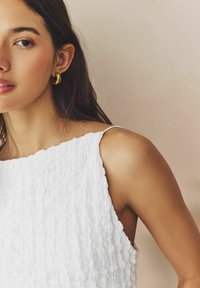 Vestito bianco con texture, scollo quadrato e spalline regolabili. La modella indossa un orecchino a cerchio dorato; lo sfondo presenta una tonalità neutra e morbida.