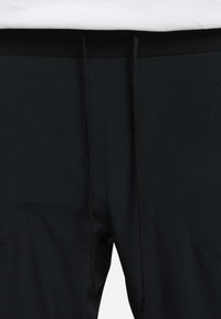 Shorts de sport noirs avec une texture lisse, présentant une taille noire et des cordons de serrage ajustables. Des poches latérales sont visibles.