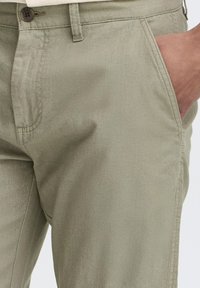 Chinos en coton vert clair avec une texture lisse, dotés d'une poche latérale et d'une fermeture à bouton marron au niveau de la taille.