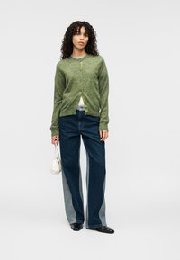 Πράσινη μάλλινη ζακέτα με κούμπωμα κουμπιών, σε συνδυασμό με σκούρο μπλε τζιν με σκίσιμο, συνοδευόμενη από μικρή λευκή τσάντα χειρός.