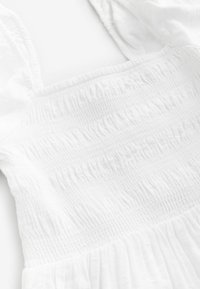 Witte jurk met een geraffineerde smock-borstvoeding en een vloeiende rok. De stof heeft een zachte textuur en een subtiele glans.