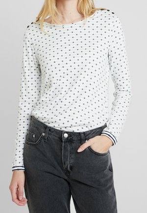 Kvinde iført en hvid langærmet top med små sorte polkaprikker og mørke højtaljede jeans, den ene hånd i lommen, foran en ensartet baggrund.