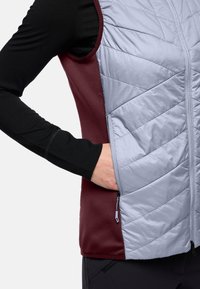 Hellblaues isoliertes Weste mit einer kastanienbraunen Seite, ausgestattet mit einer Reißverschlusstasche und einer gesteppten Textur an der Vorderseite. Schwarzes Langarmshirt darunter.