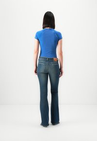 Woman standing with back to camera wearing a fitted blue short-sleeve top and flared blue jeans against a plain white background.