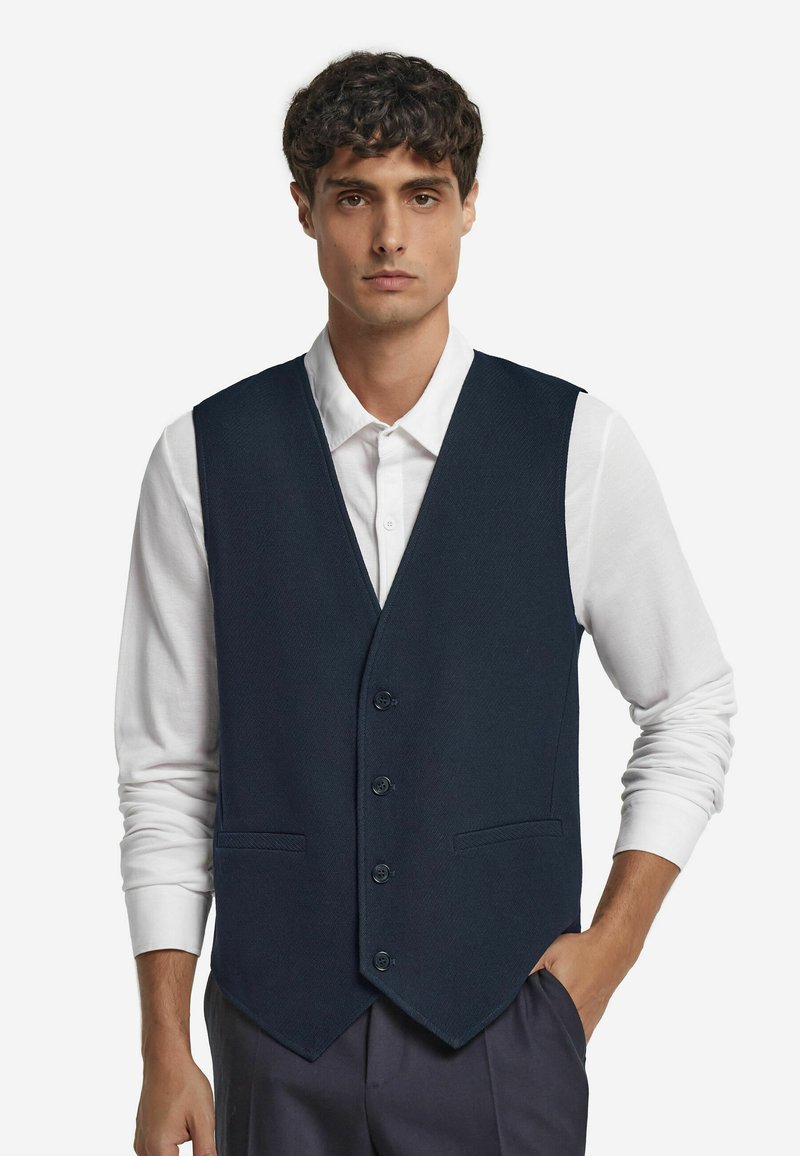 Gilet navy con tessuto testurizzato, chiusura a cinque bottoni e due tasche frontali, indossato sopra una camicia bianca con colletto.