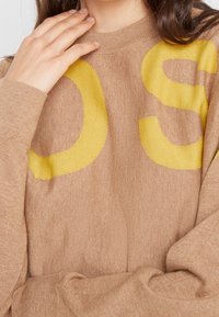 Brauner Strickpullover mit großen gelben Buchstaben "OS". Der Pullover hat einen runden Hals und lange Ärmel aus einem weichen, strukturierten Stoff.