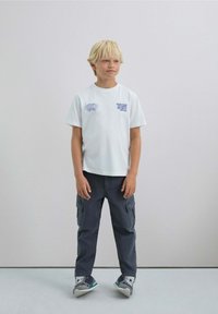 Camiseta blanca de manga corta con gráfico azul, combinada con pantalones cargo gris oscuro con bolsillos y zapatos con acentos azules.