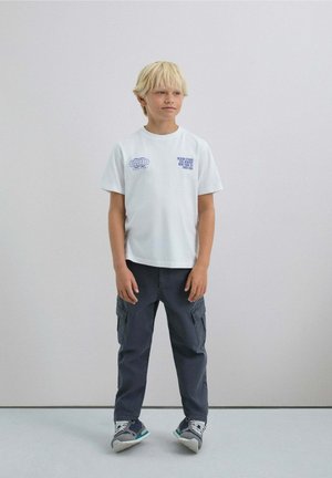 Camiseta blanca de manga corta con gráfico azul, combinada con pantalones cargo gris oscuro con bolsillos y zapatos con acentos azules.
