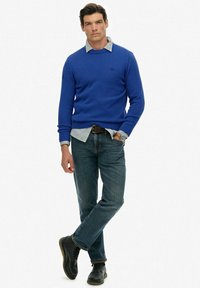 Man in blauw sweater over een grijs shirt, blauwe jeans, zwarte schoenen en een polshorloge, staand met één been over het andere gekruist.