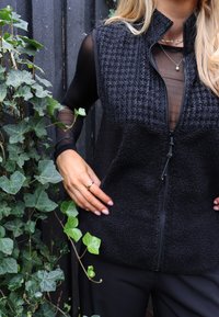 Melna veste ar rāvējslēdzēju un teksturētu priekšpusi, izgatavota no flīsa materiāla, uzvilkta virs caurspīdīgas melnas garām piedurknēm blūzes. Fonā zaļš efejas augs.