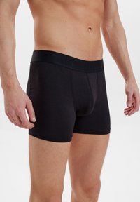 Schwarze Baumwoll-Boxershorts mit einem weichen Bündchen, das ein dezentes Logo zeigt. Fitted-Design und glatte Textur, präsentiert an einem männlichen Modell.