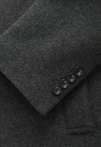 Manga de abrigo de lana gris oscuro con un acabado texturizado. Presenta cuatro botones negros en el puño y un pequeño bolsillo abierto.
