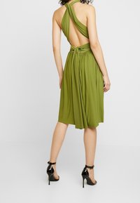 Robe sans manches vert olive avec un design noué dans le dos, tissu fluide, et ourlet mi-long, associée à des escarpins noirs à talons aiguilles.