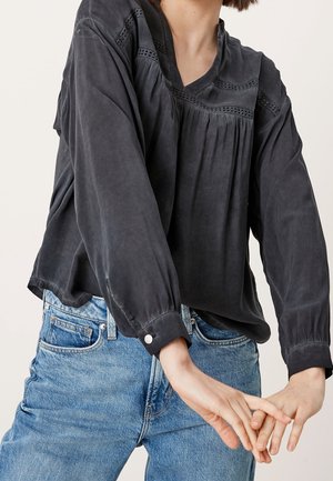 Personne portant un chemisier noir ample à manches longues avec des détails en dentelle et un jean en denim bleu, les mains jointes devant.