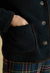 Giacca in pile blu scuro con piping marrone, dotata di tasca frontale e bottoni arrotondati. Indossata sopra pantaloni a quadri rossi e verdi.