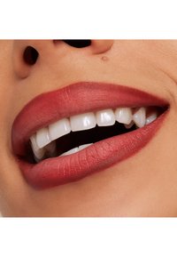 Zbliżenie na usta z pełnymi wargami pomalowanymi matową, czerwonawobrązową szminką oraz białymi zębami. Skóra ma gładką teksturę w naturalnych odcieniach.