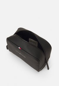 Sorte tekstureret læder toilettaske med åbent lynlås, der viser indre label, Tommy Hilfiger logo og signaturflag på fronten.