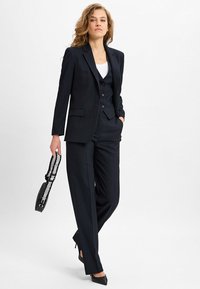 Vrouw die loopt in een marineblauw gestreept pak met vest, witte top, zwarte hakken en een zwarte handtas met een patroon op de schouderband.