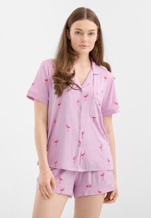 Nuori nainen pukeutuneena liilanväriseen lyhythihaiseen napitettavaan paitaan, jossa on punainen flamingokuvio, sekä samaa sarjaa oleviin shortseihin, seisoo valkoista taustaa vasten.