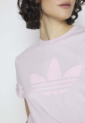 Kobieta z kręconymi ciemnymi włosami, ubrana w jasnoróżowy t-shirt z dużym logo Adidas trefoil na przodzie.