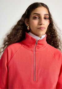 Felpa corallo con zip e collo alto, caratterizzata da una texture a coste e maniche raglan, abbinata a una camicia bianca con colletto sotto.