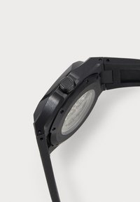 Schwarze Uhr mit achteckigem Gehäuse, strukturiertem Finish, sichtbarem Uhrwerk durch einen transparenten Boden und sechseckiger Krone.