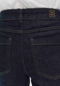 Donkere spijkerbroek met gele stiksels, voorzien van een subtiel logo op de tailleband en twee achterzakken. Soepele textuur en een aangesloten pasvorm.