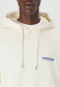 Hoodie couleur crème avec cordons, arborant un logo bleu sur la poitrine gauche. Fabriqué à partir d'un tissu doux, il possède une coupe décontractée et un design à enfiler.