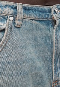 Κοντινό πλάνο μπλε τζιν, που δείχνει τη μέση, το θηλάκι ζώνης, το μεταλλικό κουμπί, το ριβέτ και τις λεπτομέρειες ραφής.