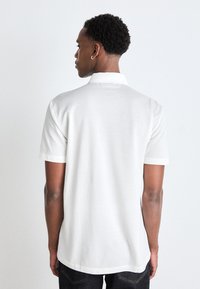 Bílá polo tričko s krátkým rukávem, rovného střihu, měkké textury, s ohrnutými manžetami a límcem. Jednoduchý design bez vzorů nebo akcentů.